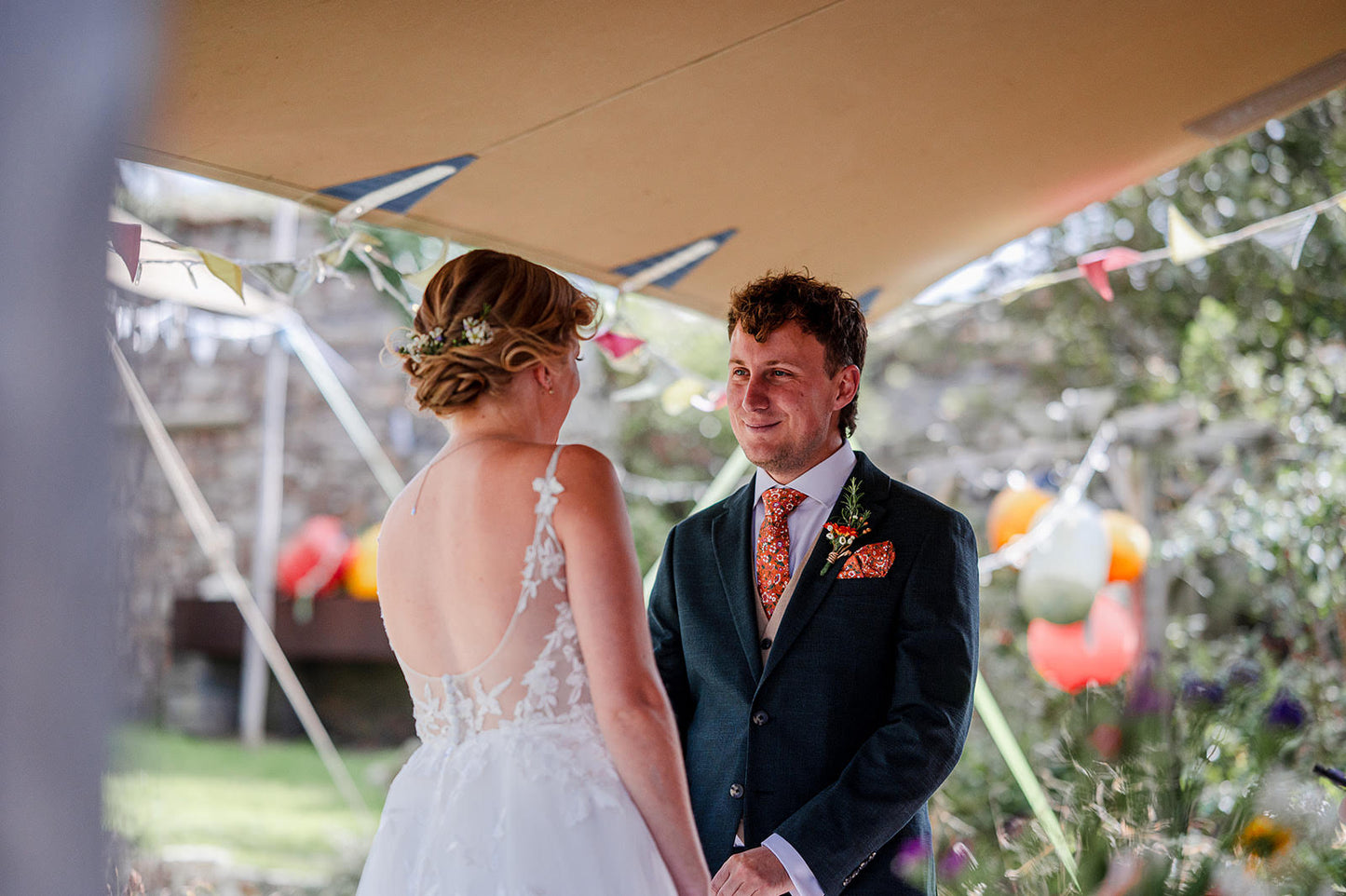 A bride and groom holding hands. She is wearing a white lace gown and the groom has a burnt orange floral tie and pocket square paired with a navy suit.