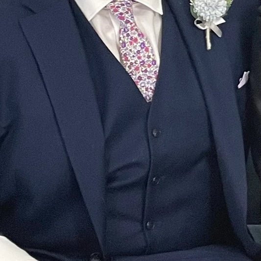 A lavender floral cotton men's tie with purple flowers and green accents matched with a navy suit. The ties are hand made by Daisy and Oak Studio and come gift wrapped so make ideal men's gifts and grooms and groomsmen ties.