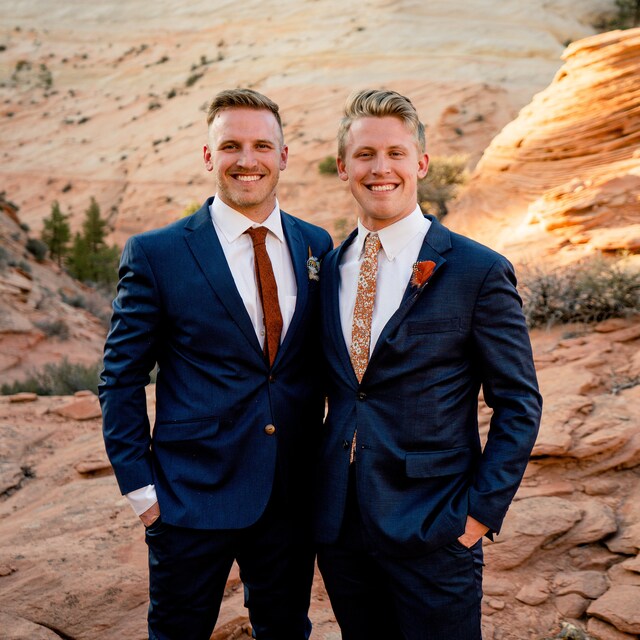 A groom and groomsman wearing an orange floral tie and burnt orange pocket square paired with a navy suit.