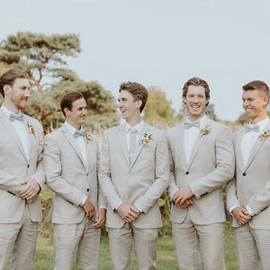 A groom and groomsmen at a wedding wearing a light grey, handmade herringbone wool bow tie and mens necktie. The men are wearing light beige suits paired with grey ties and bow ties.
